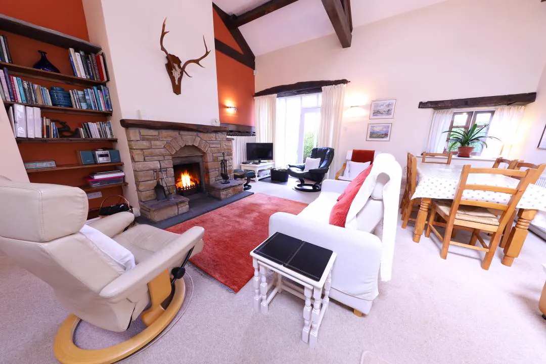 The open-plan living room with the fire lit, exposed roof beams above and a dining table by the window
